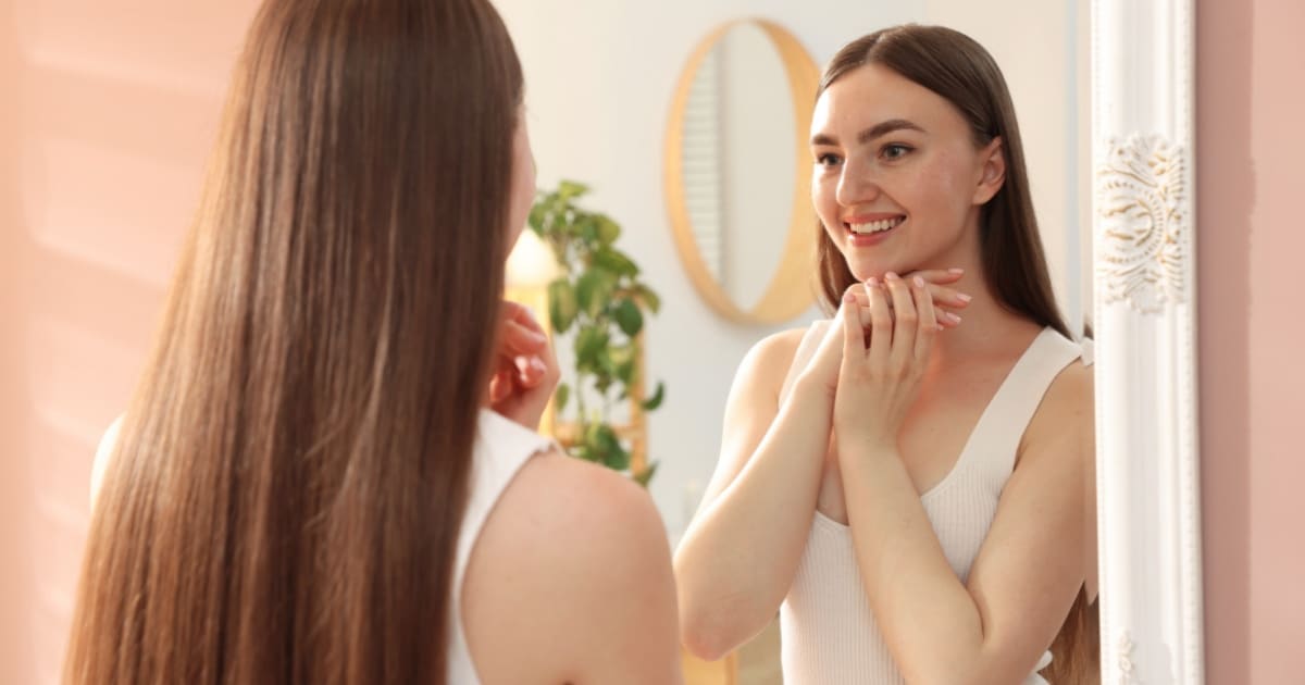 A young woman smiling at her reflection in a mirror, standing in a softly lit room with light pink walls and a plant in the background.