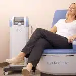 A woman sits comfortably in a blue medical chair labeled “BTL EMSELLA,” smiling with her eyes closed as she receives a non-invasive pelvic floor treatment in a clean clinical room, with the treatment device positioned beside the chair.