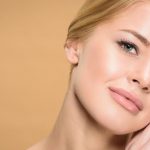Close-up of a woman with clear skin gently touching her face against a soft beige background.
